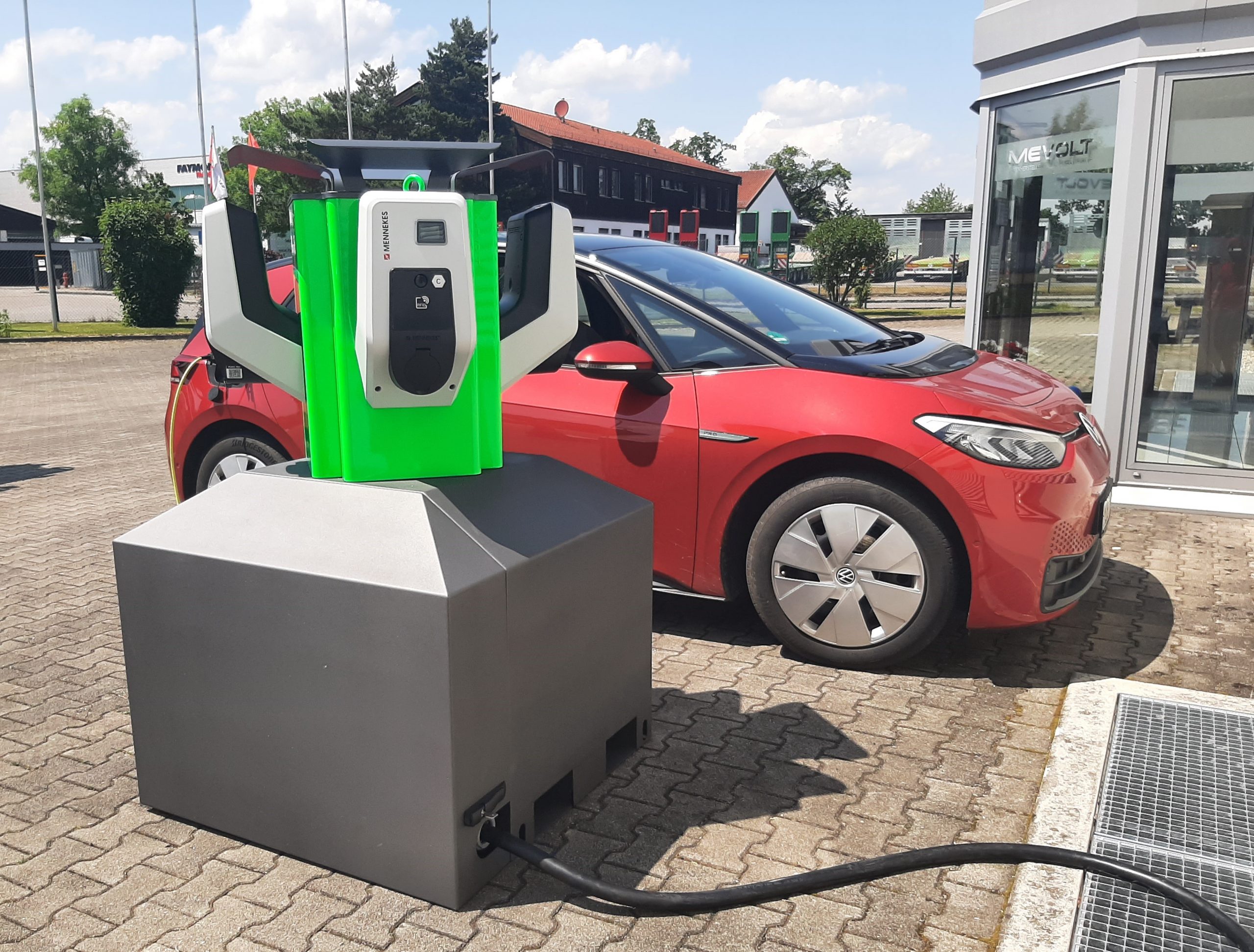Der mobile Schnelllader "move charger" von meevolt steht auf einem Parkplatz vor einem roten VW ID.3 Elektroauto. Die kompakte Ladesäule mit grünem Gehäuse und weißen Griffarmen ist auf einem grauen Sockel montiert und über ein Kabel mit Strom versorgt. Im Hintergrund ist ein moderner Gebäudekomplex zu sehen.